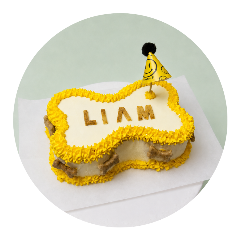Torta en forma de hueso de perro color blanca con decoraciones en amarillo y el nombre "liam" hecho en galletitas para perros